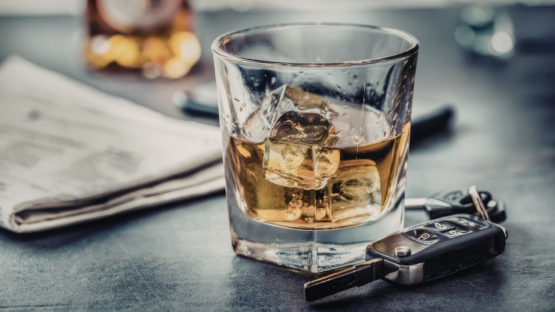 Un vaso de alcohol con hielos sobre una mesa y a su lado aparece un periódico y unas llaves de un vehículo.